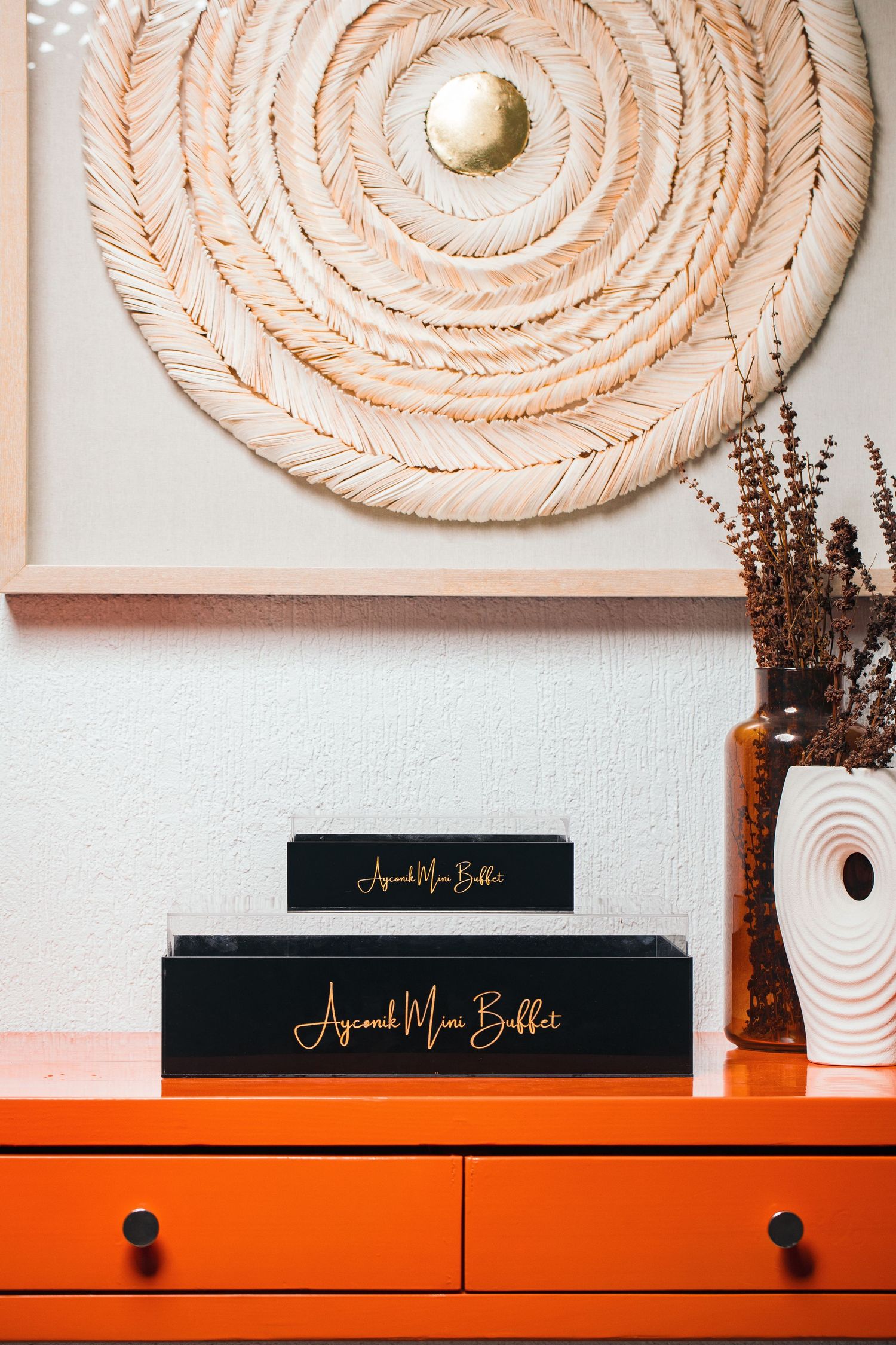 Circular woven wall art above an orange dresser with black boxes and dried flowers in a vase.