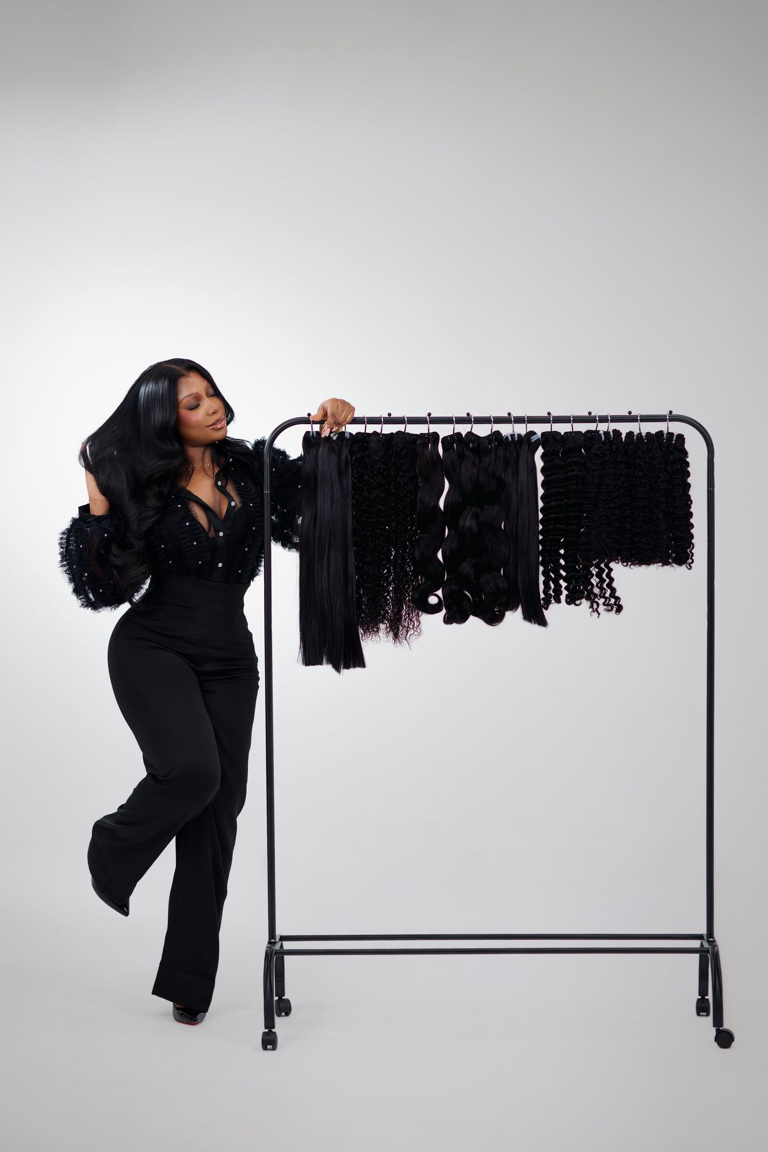 Woman in black outfit posing beside a clothing rack displaying various black hair extensions and weaves.