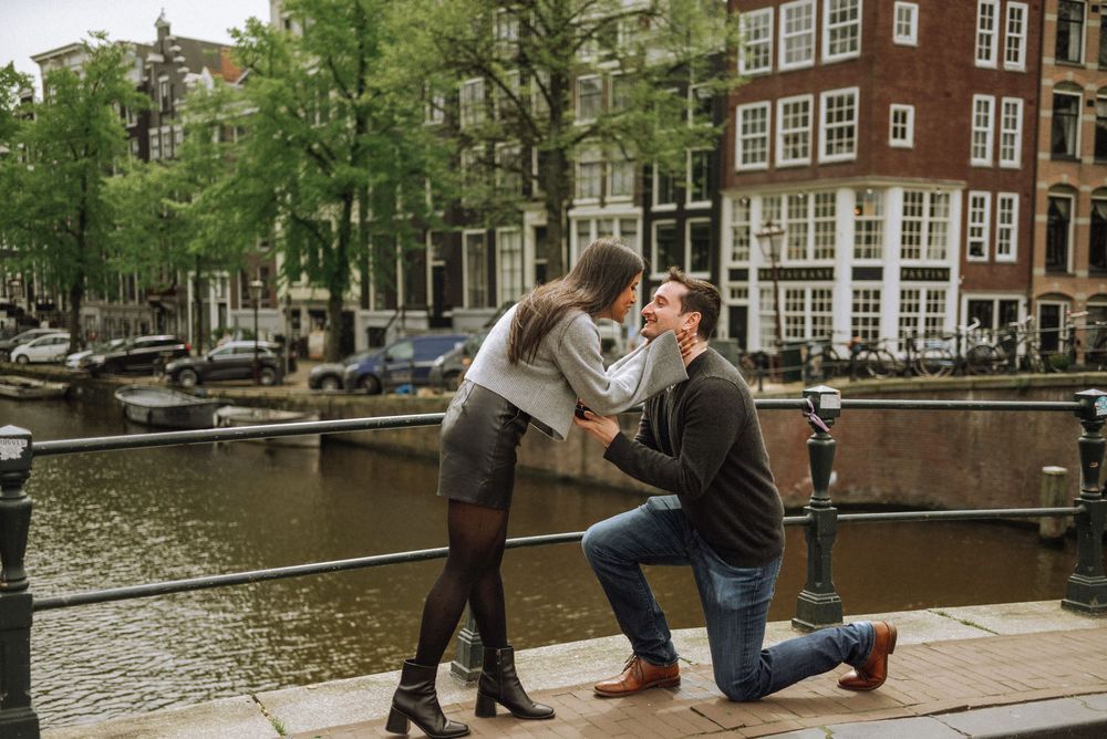 A romantic proposal scene along an Amsterdam canal with historic buildings in the background.