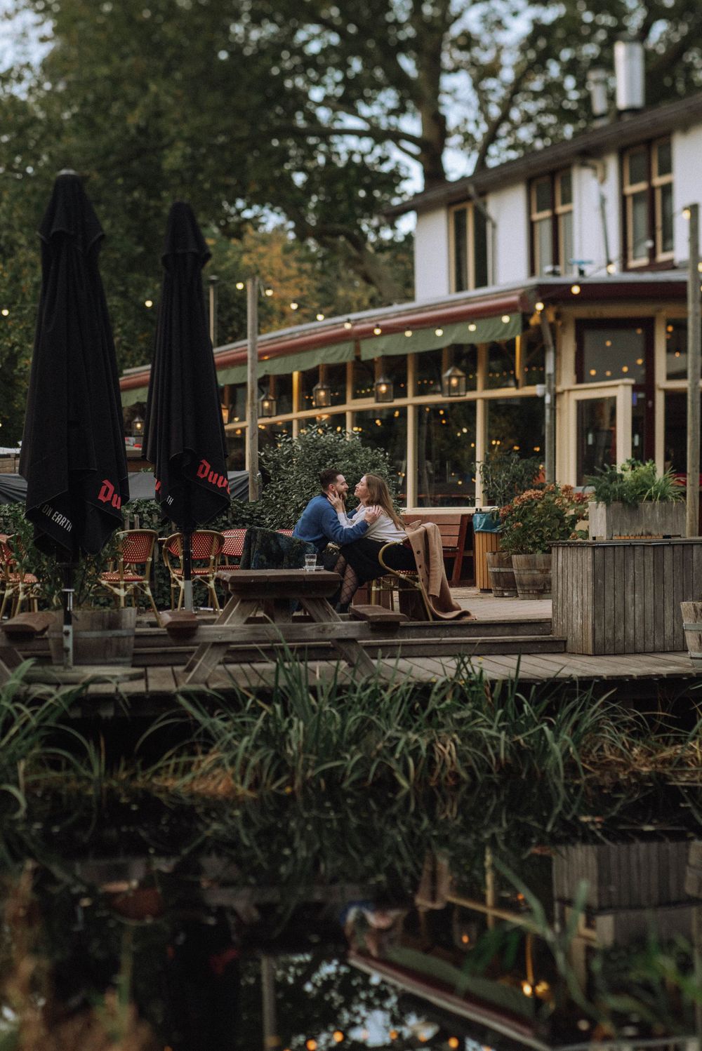 A romantic restaurant patio setting with string lights reflected in water at dusk.