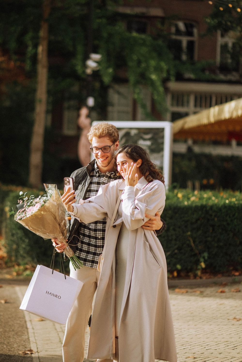 A couple shares a joyful moment while holding shopping bags and flowers on a city sidewalk.