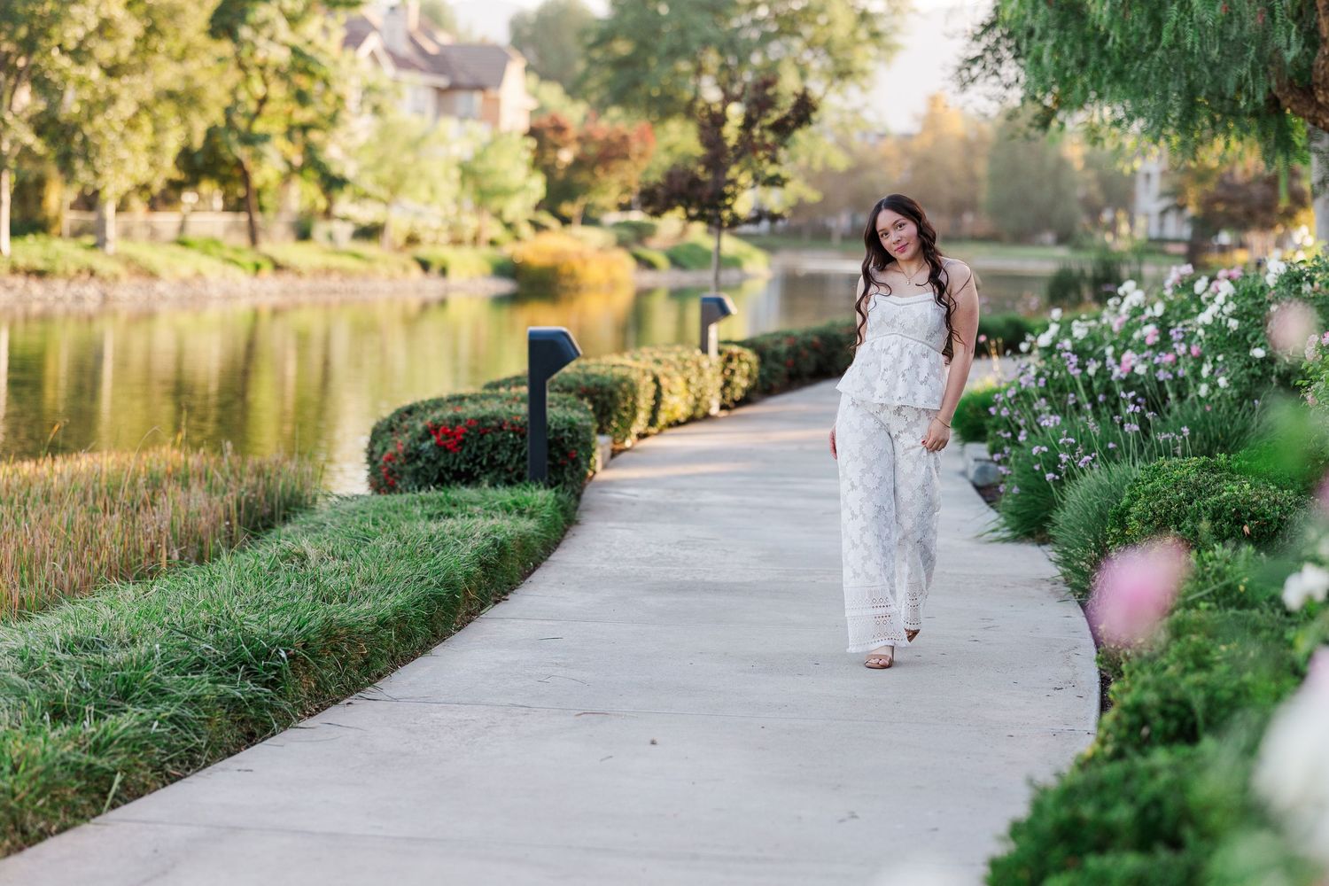 A person in a white lace outfit walks along a scenic lakeside path surrounded by lush greenery and flowers.