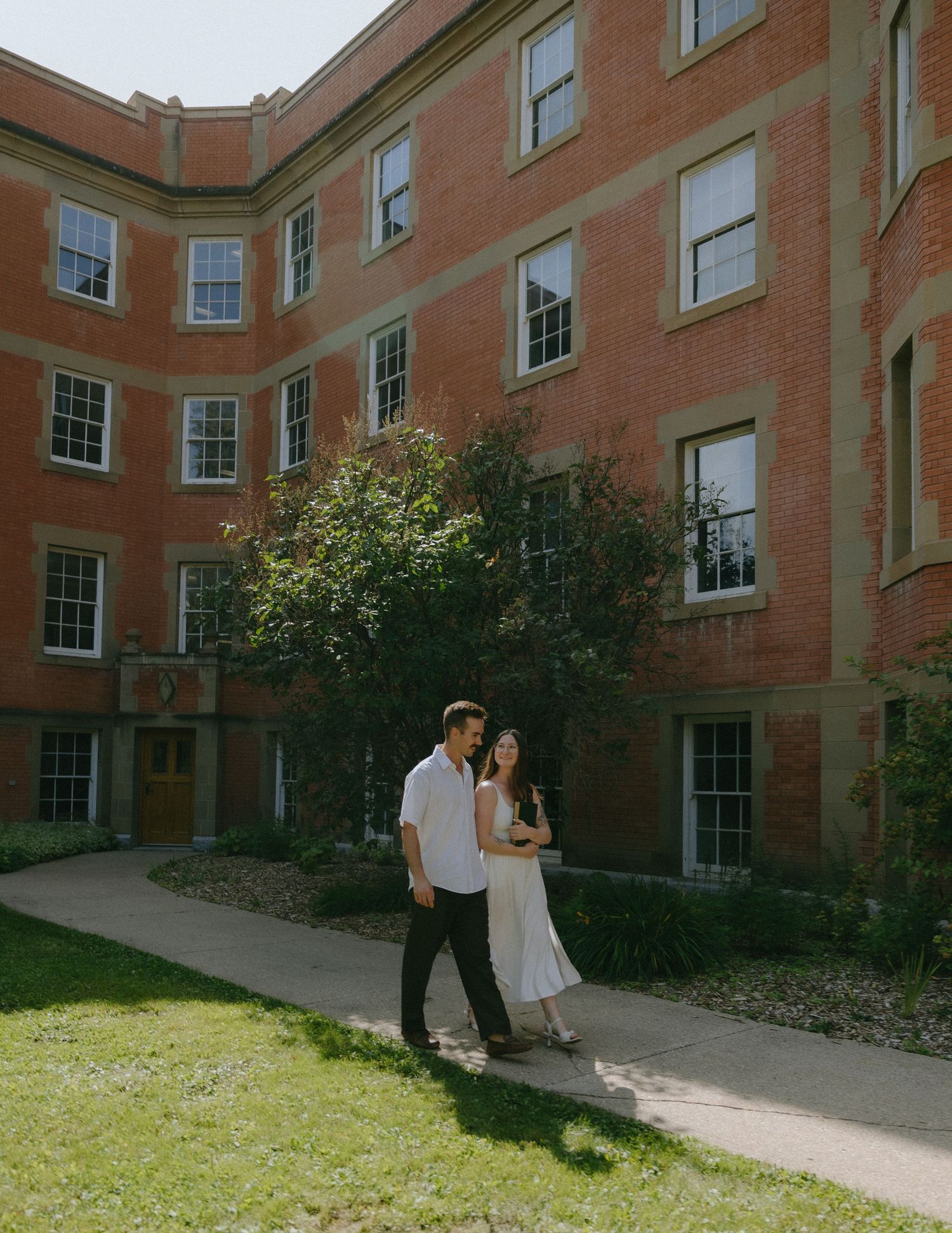 A couple shares a romantic moment together outside a historic red brick building on a sunny day.