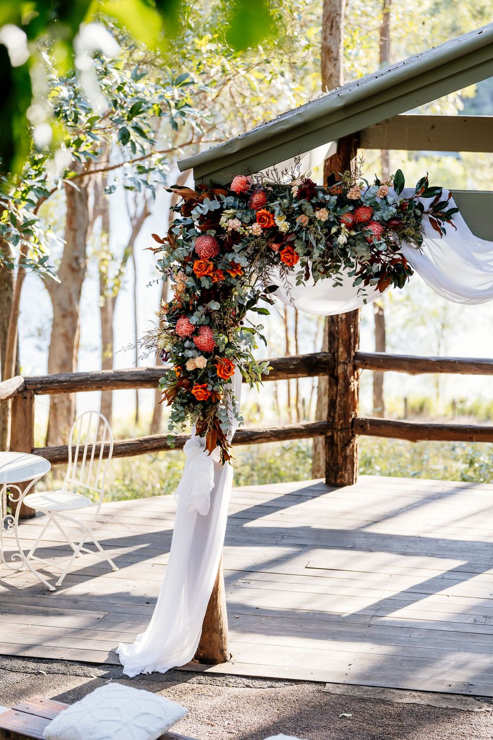 Rustic outdoor wedding ceremony arch decorated with red and white flowers and flowing fabric against a sunlit forest backdrop.