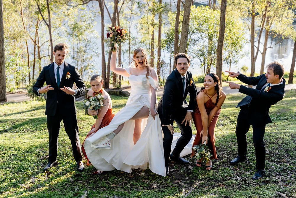 Wedding party poses playfully in a sunlit forest setting with trees and natural greenery in the background.