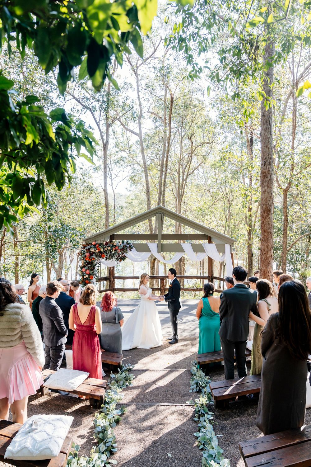 An outdoor wedding ceremony takes place under a rustic wooden pavilion surrounded by tall trees in a forest setting.