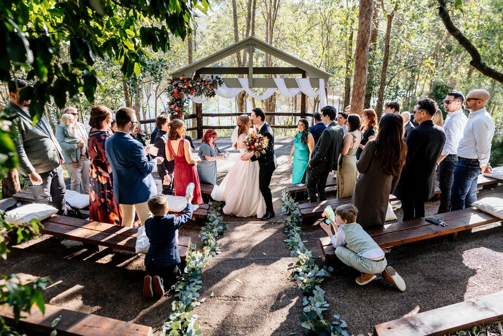 Wedding guests gather under a rustic wooden pavilion surrounded by lush greenery and natural light.
