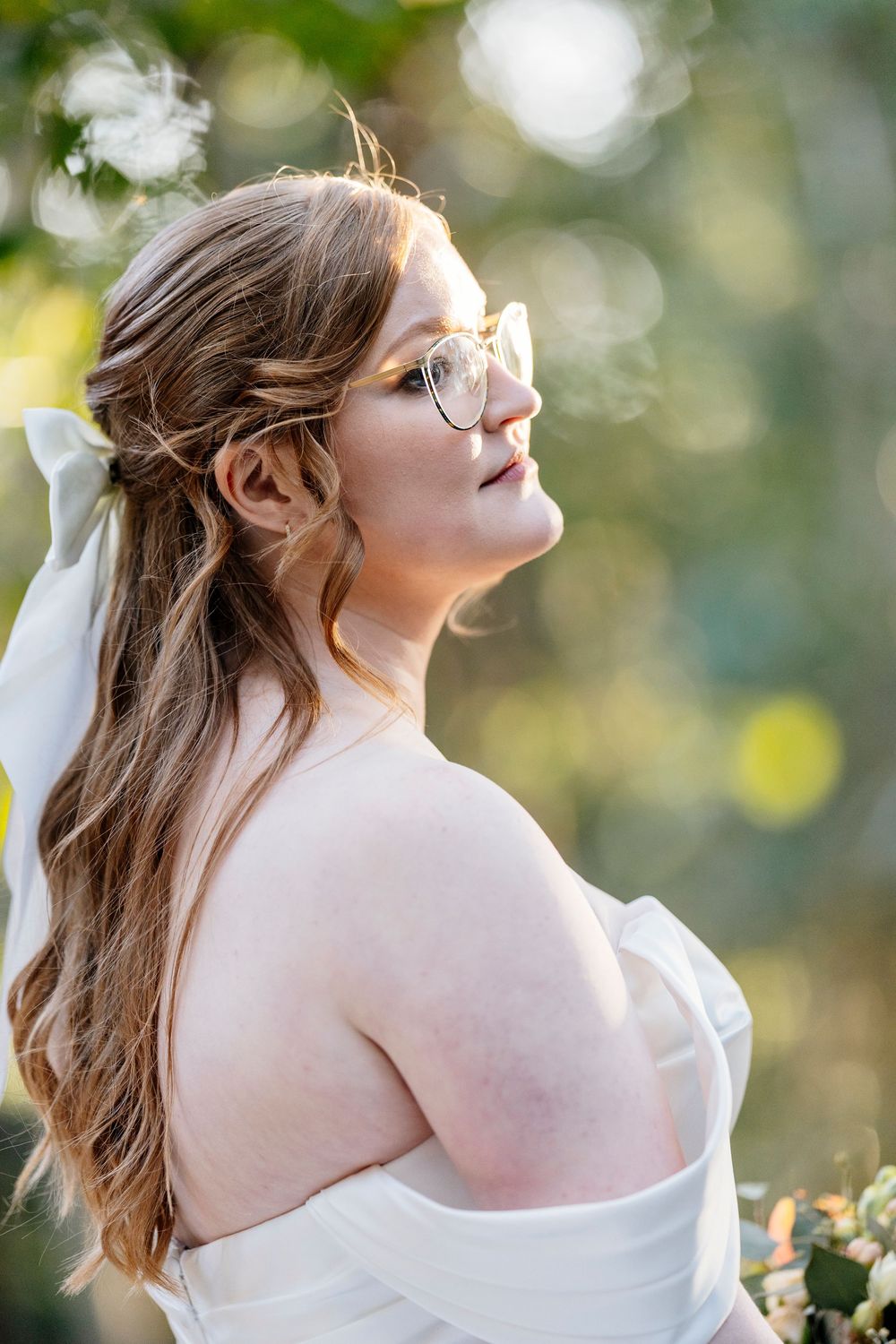 A bride in a white dress and glasses gazes upward in soft natural lighting with bokeh effect in the background.