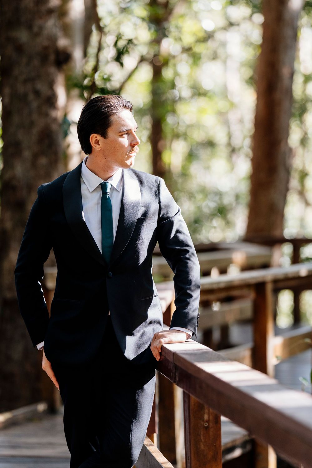 A person in a dark suit and tie stands on a wooden deck surrounded by sunlit trees and forest.