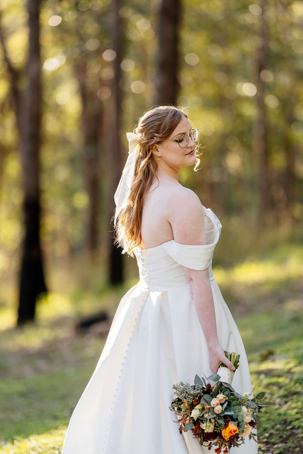 A bride in an off-shoulder white wedding gown holds an autumn bouquet while standing in a sunlit forest setting.