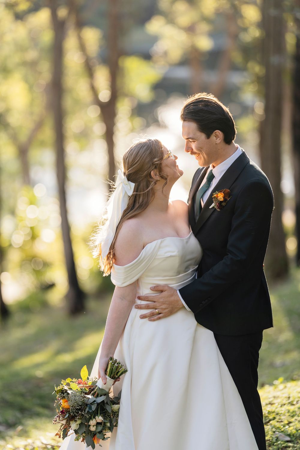 A romantic wedding couple embraces in a sun-dappled forest setting with golden afternoon light filtering through trees.