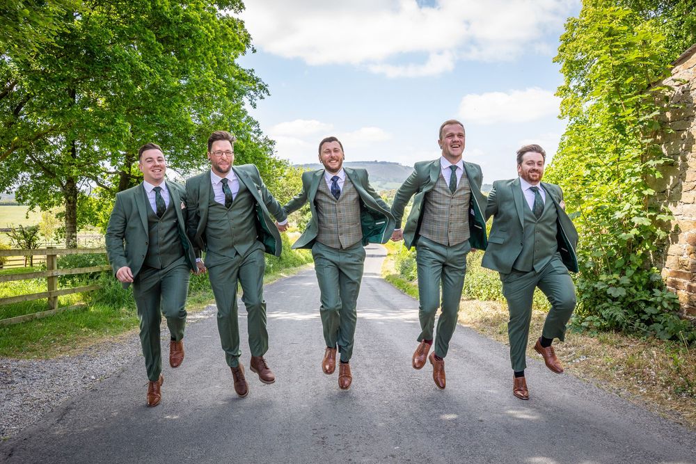 Five groomsmen in matching green suits jump joyfully together on a sunny country road lined with trees.