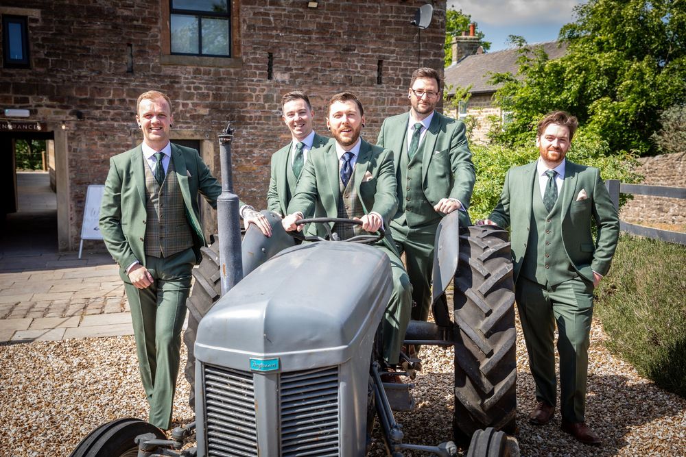 Five groomsmen in matching green suits pose confidently around a vintage tractor outside a rustic barn venue.