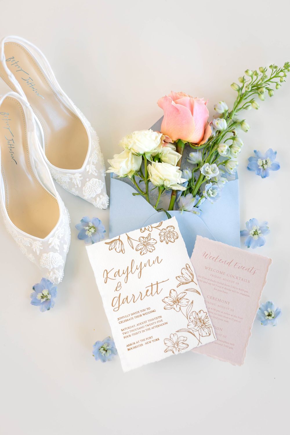 Wedding details flatlay with lace shoes, floral invitation, and pink and blue flowers.