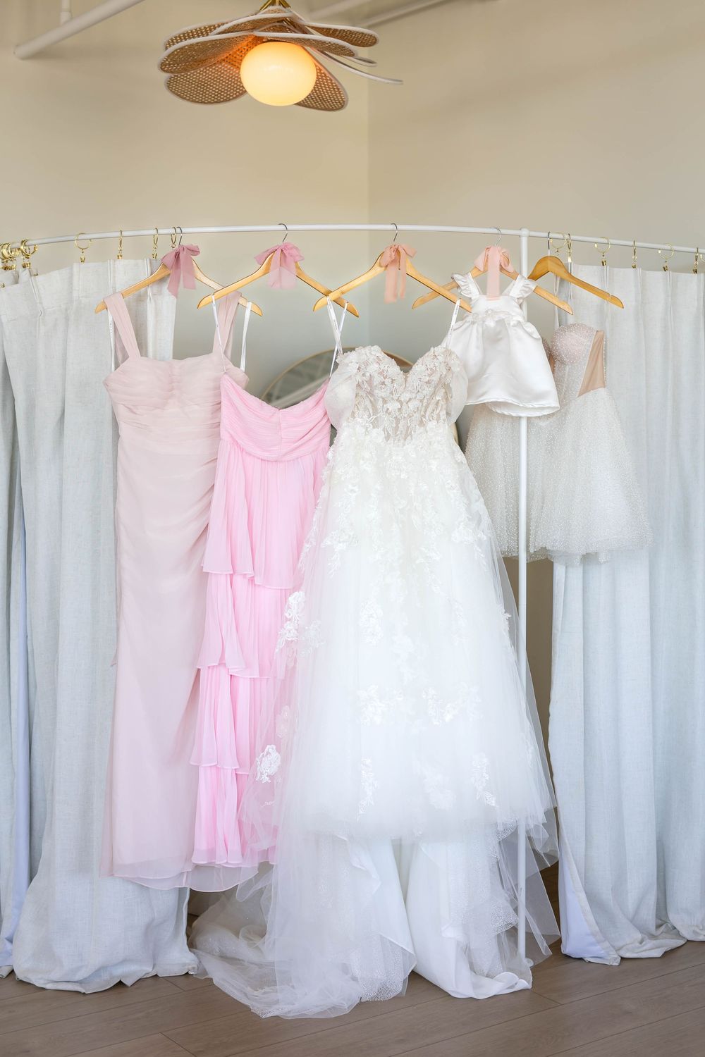 Wedding dresses and pink bridesmaid gowns hanging on a clothing rack against white curtains.