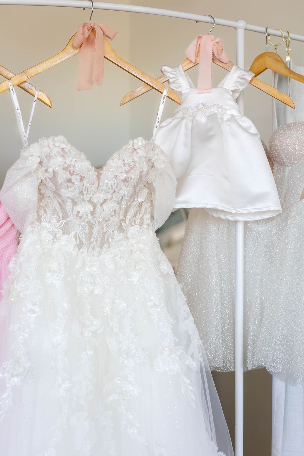 Wedding dress and flower girl dress hanging on wooden hangers with pink and white details.