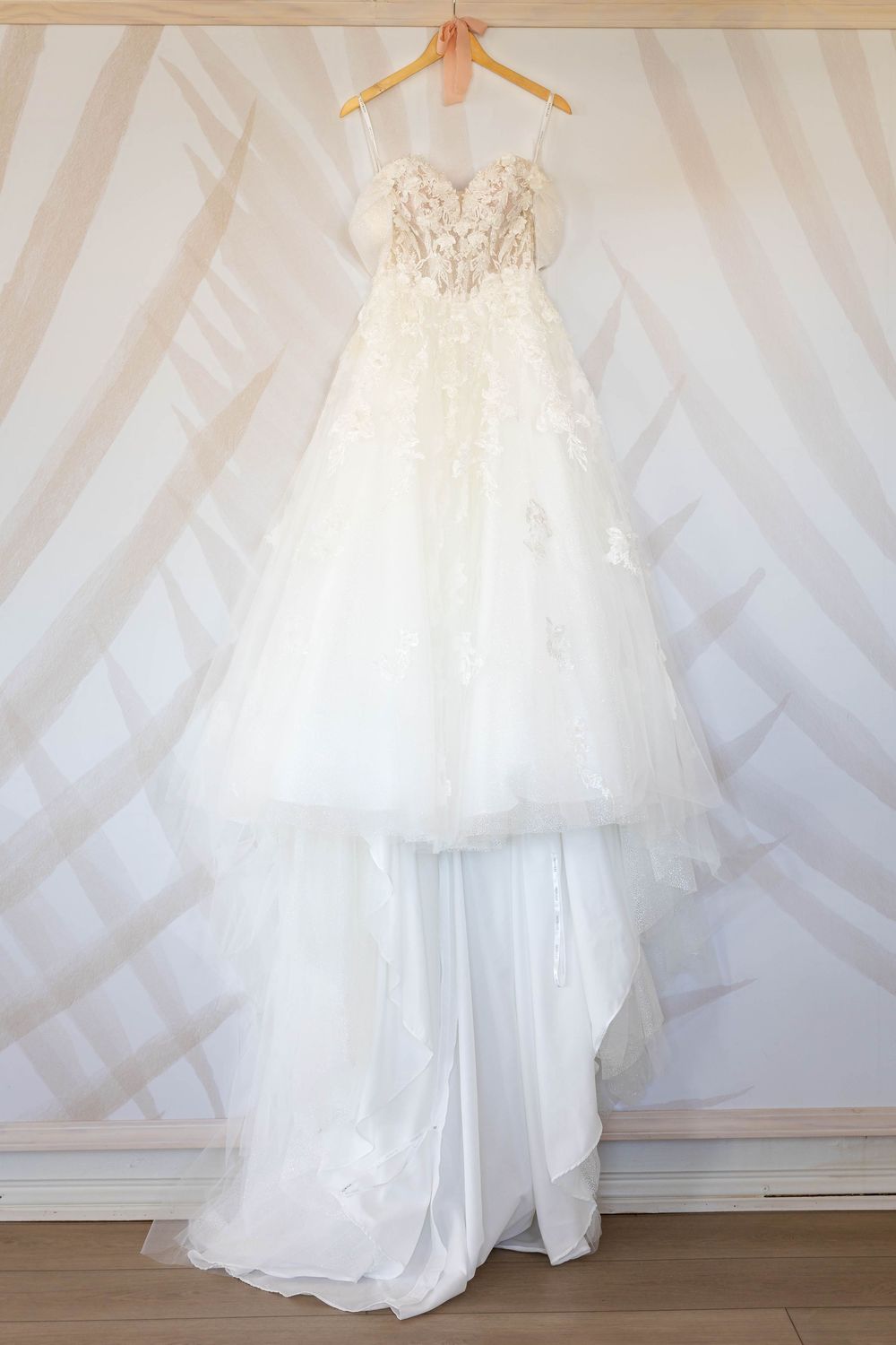 A white wedding dress with delicate lace details and flowing tulle skirt displayed on a hanger against a textured wall.