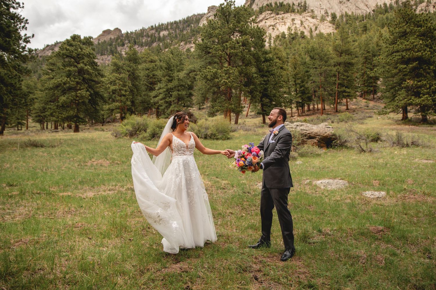 A couple in formal wedding attire poses in a scenic mountain meadow surrounded by pine trees and natural terrain.