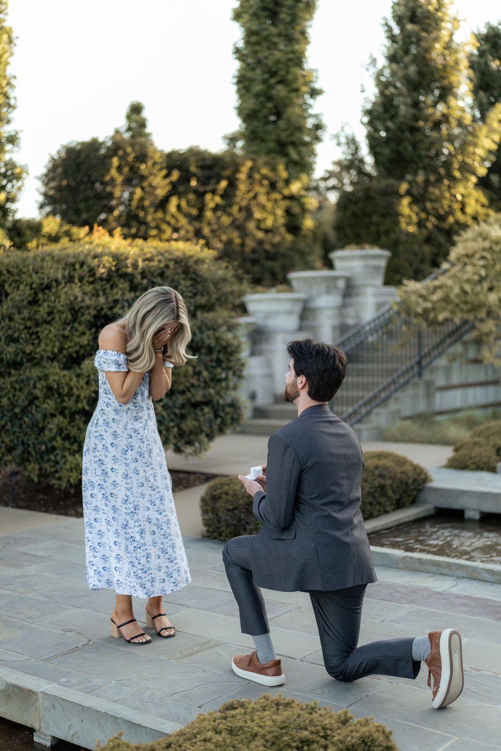Man kneeling on one knee proposing to woman in floral dress in elegant garden setting.
