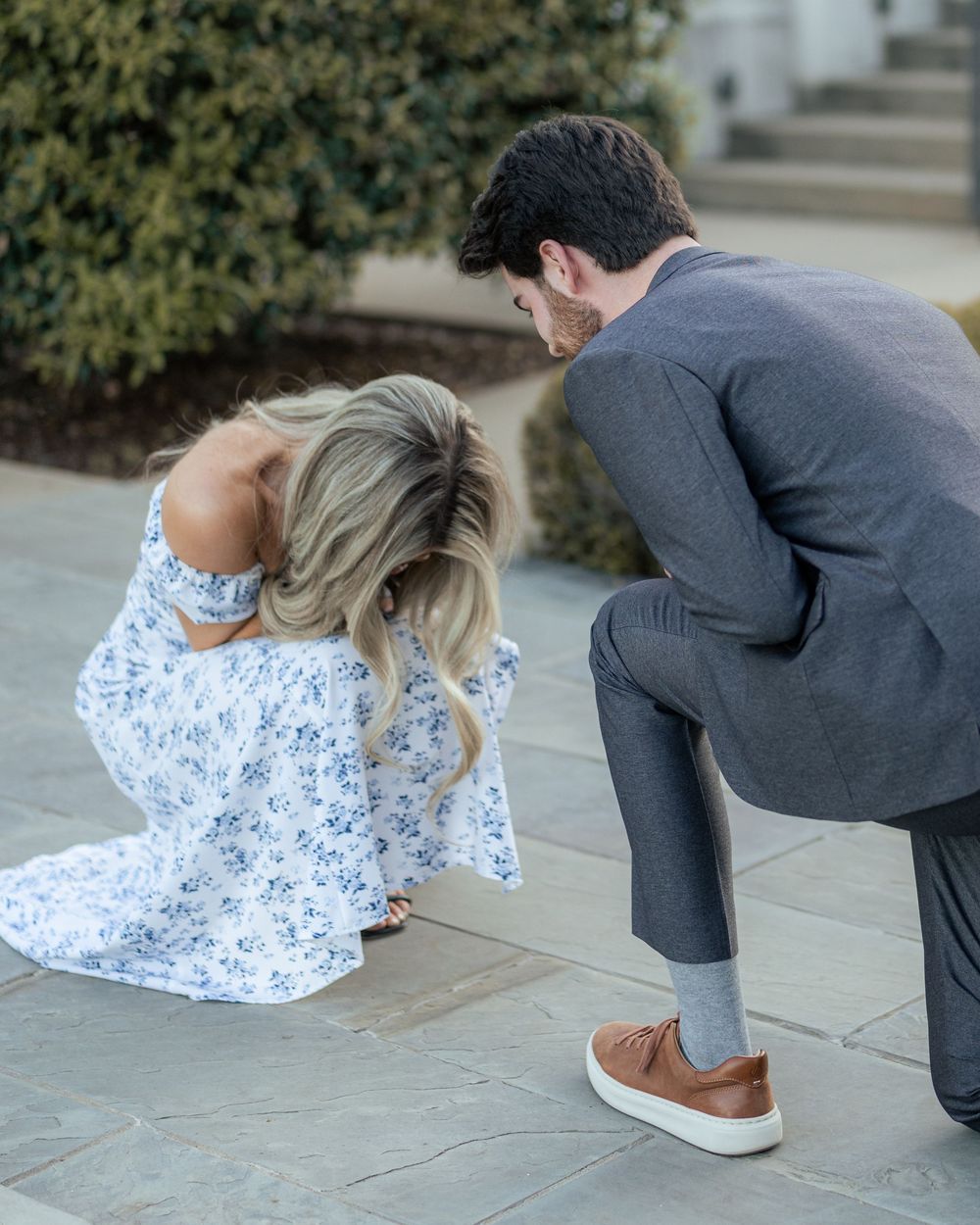 Man kneeling in gray suit proposing to blonde woman in floral dress on stone patio outdoors.
