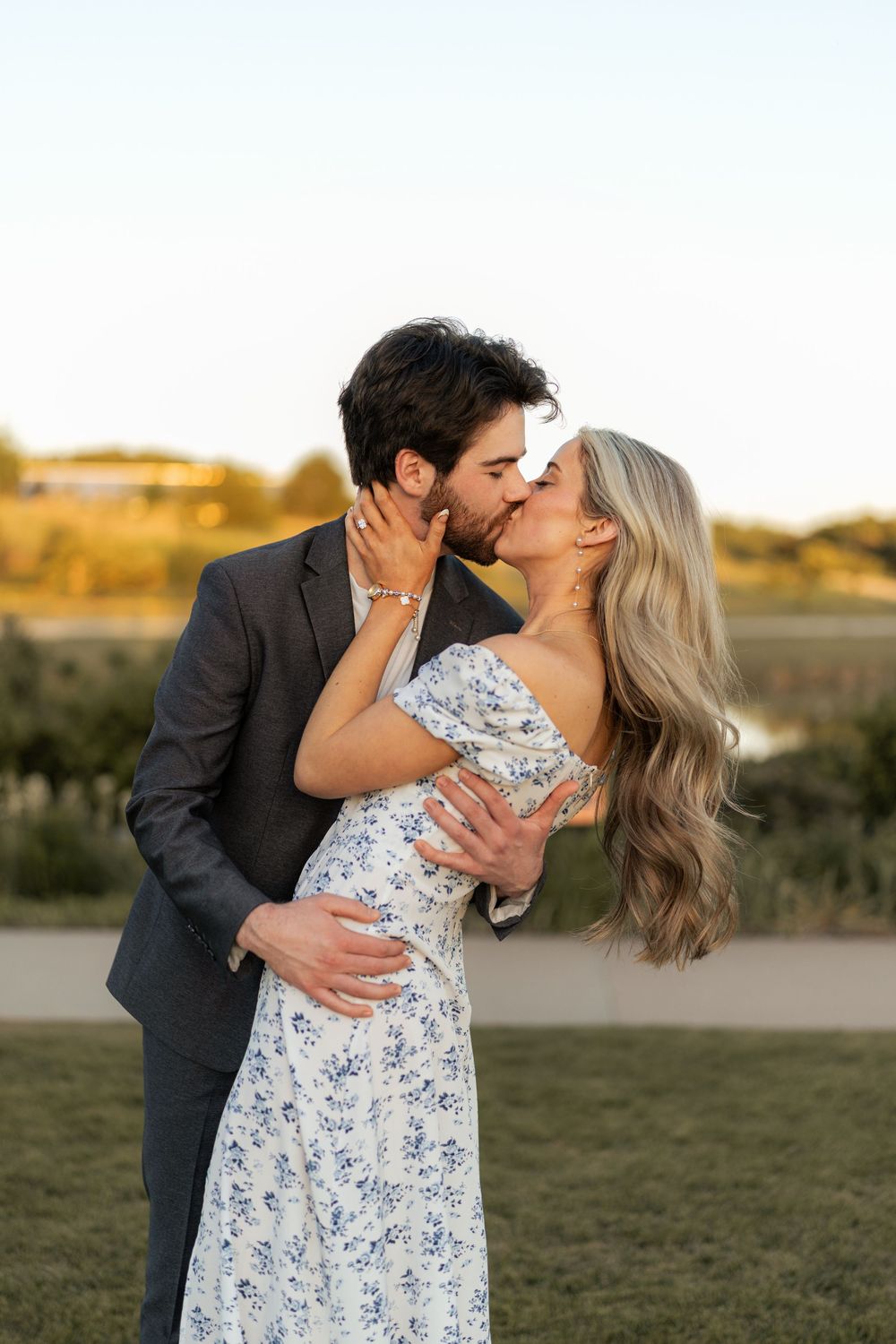 Couple sharing a romantic kiss outdoors at golden hour, man in gray suit embracing woman in floral dress.