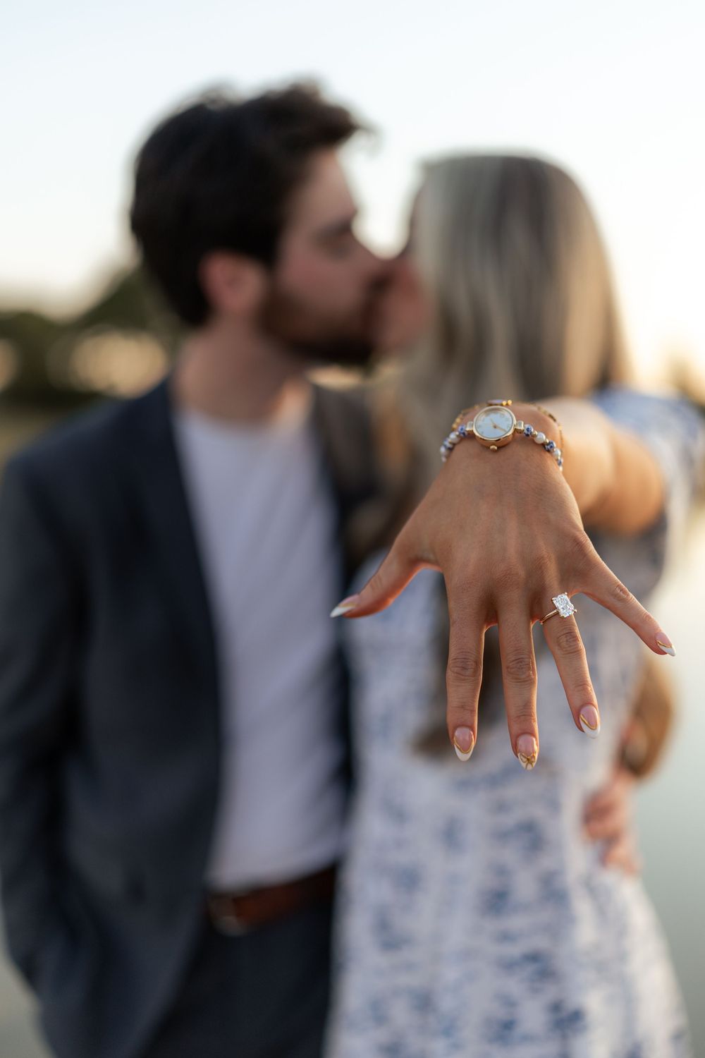 Engaged couple kissing outdoors while woman displays diamond engagement ring in foreground at sunset.