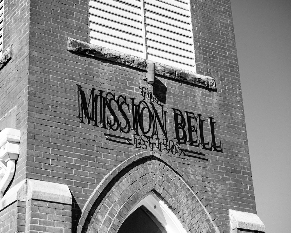 Black and white photo of The Mission Bell brick building established 1907, featuring arched windows and classic signage.