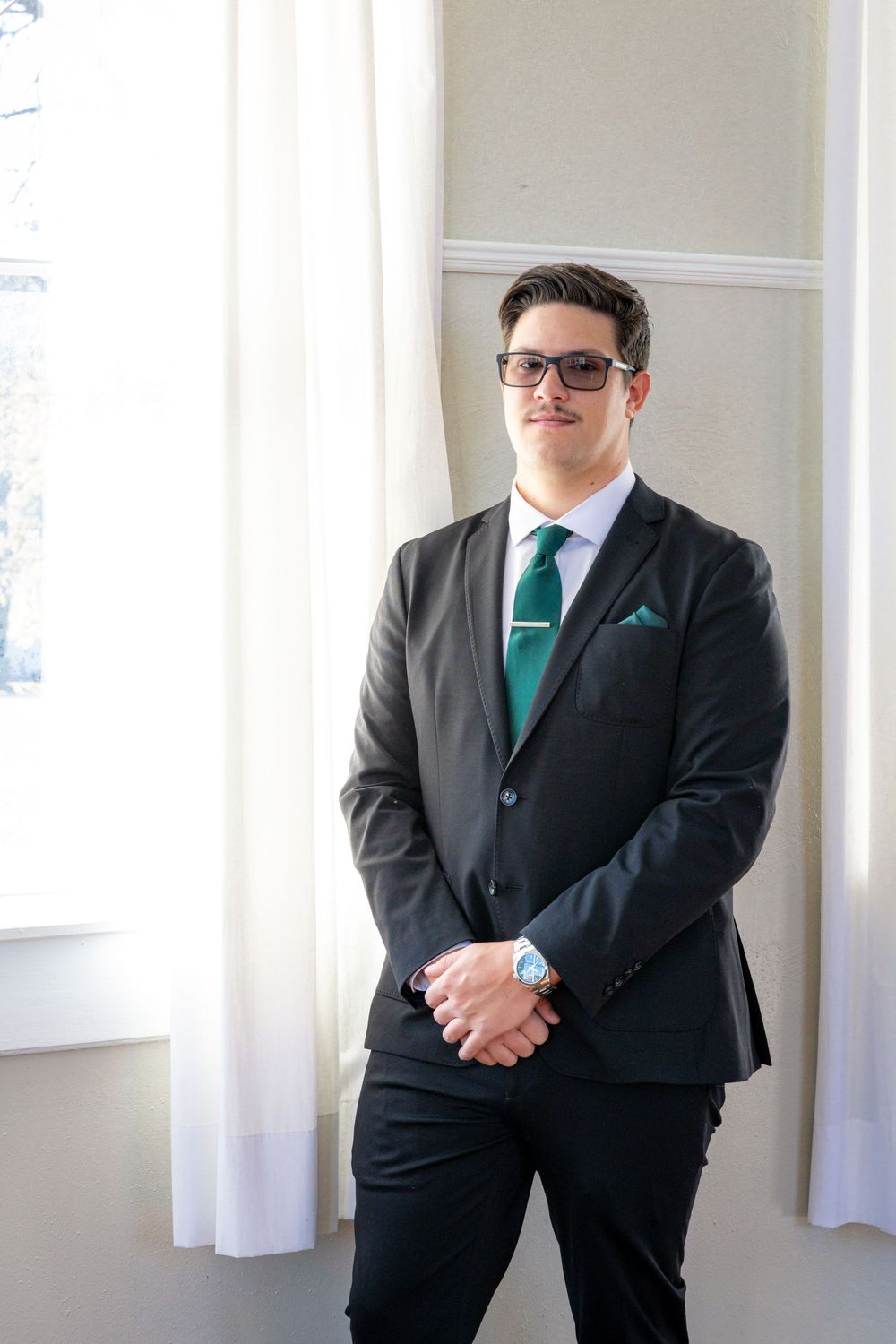 Young professional man in dark suit, green tie, pocket square, and glasses standing confidently near a bright window.