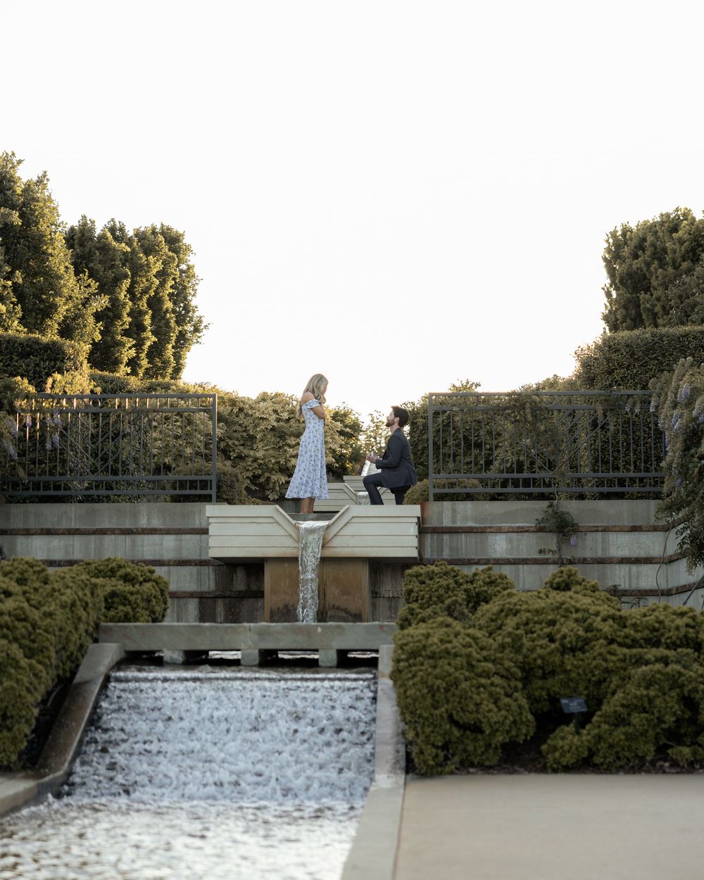 A man proposes on one knee to a woman on a garden terrace with a waterfall backdrop.