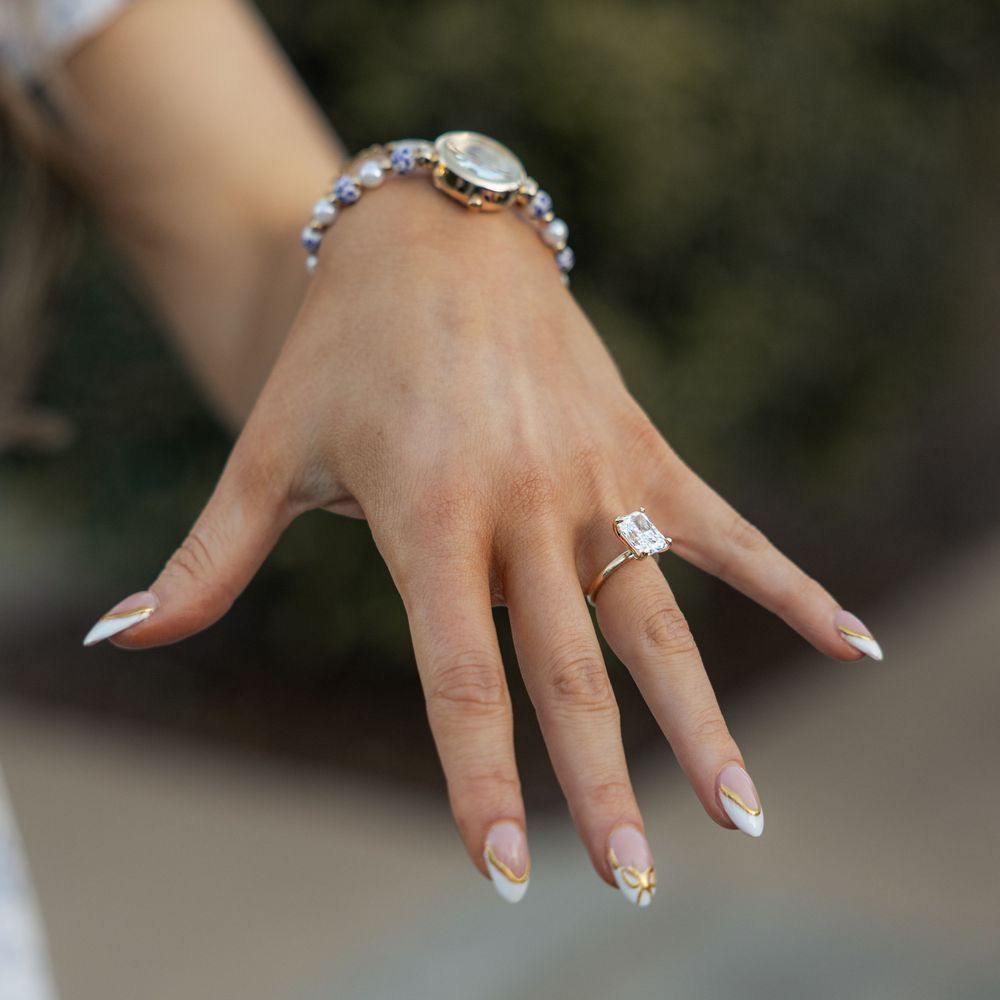 Close-up of a womans hand with elegant French tip nails, a diamond engagement ring, and a delicate beaded bracelet.