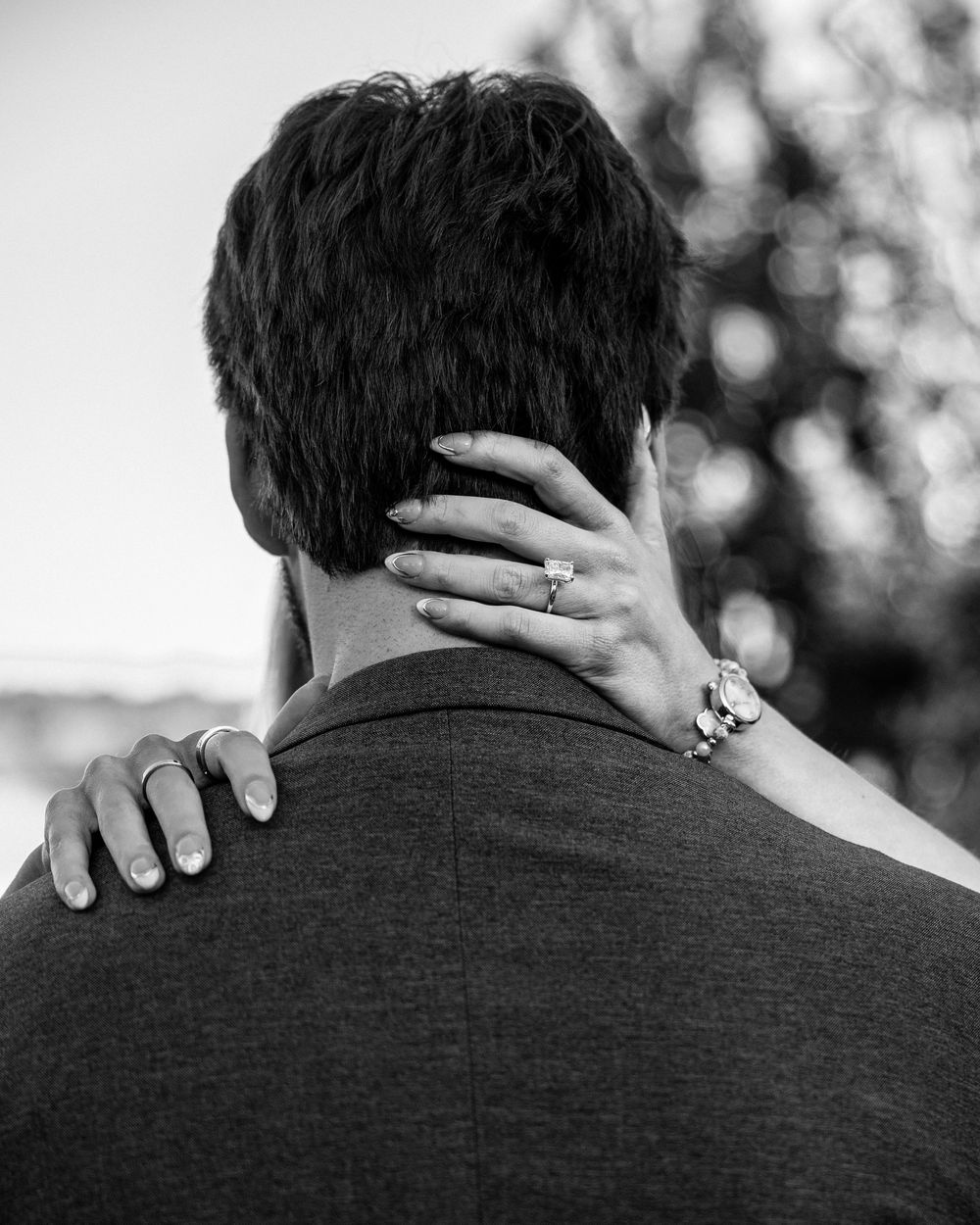 Black and white photo of a couple embracing, woman's hand with engagement ring resting on man's neck.