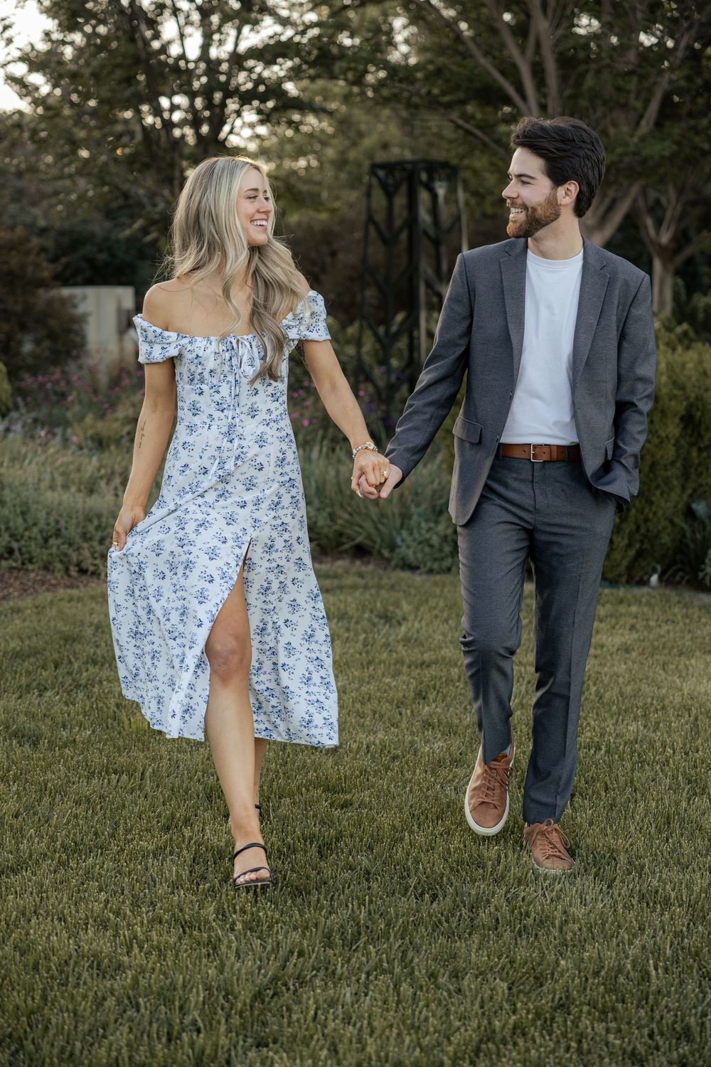 Happy couple holding hands walking in a garden, woman in floral dress, man in gray suit.