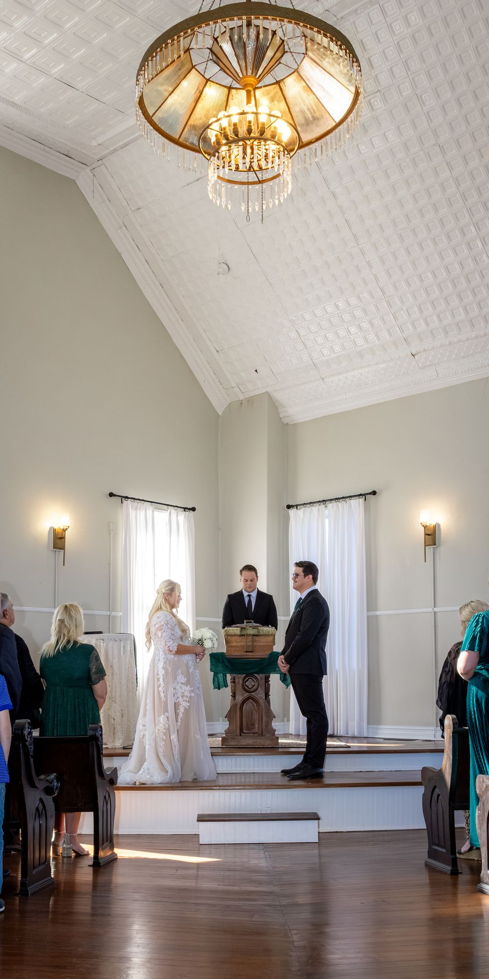 Elegant wedding ceremony in a chapel with vaulted ceiling, chandelier, bride in lace gown, groom in tuxedo at altar.