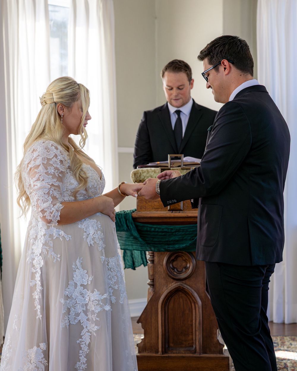 Bride and groom exchange rings at altar during indoor wedding ceremony with officiant standing behind wooden podium.