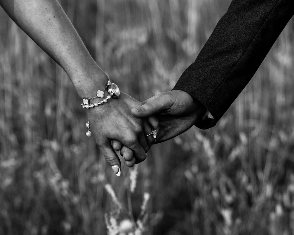 Black and white photo of a couple holding hands, with the woman wearing a charm bracelet in a field.