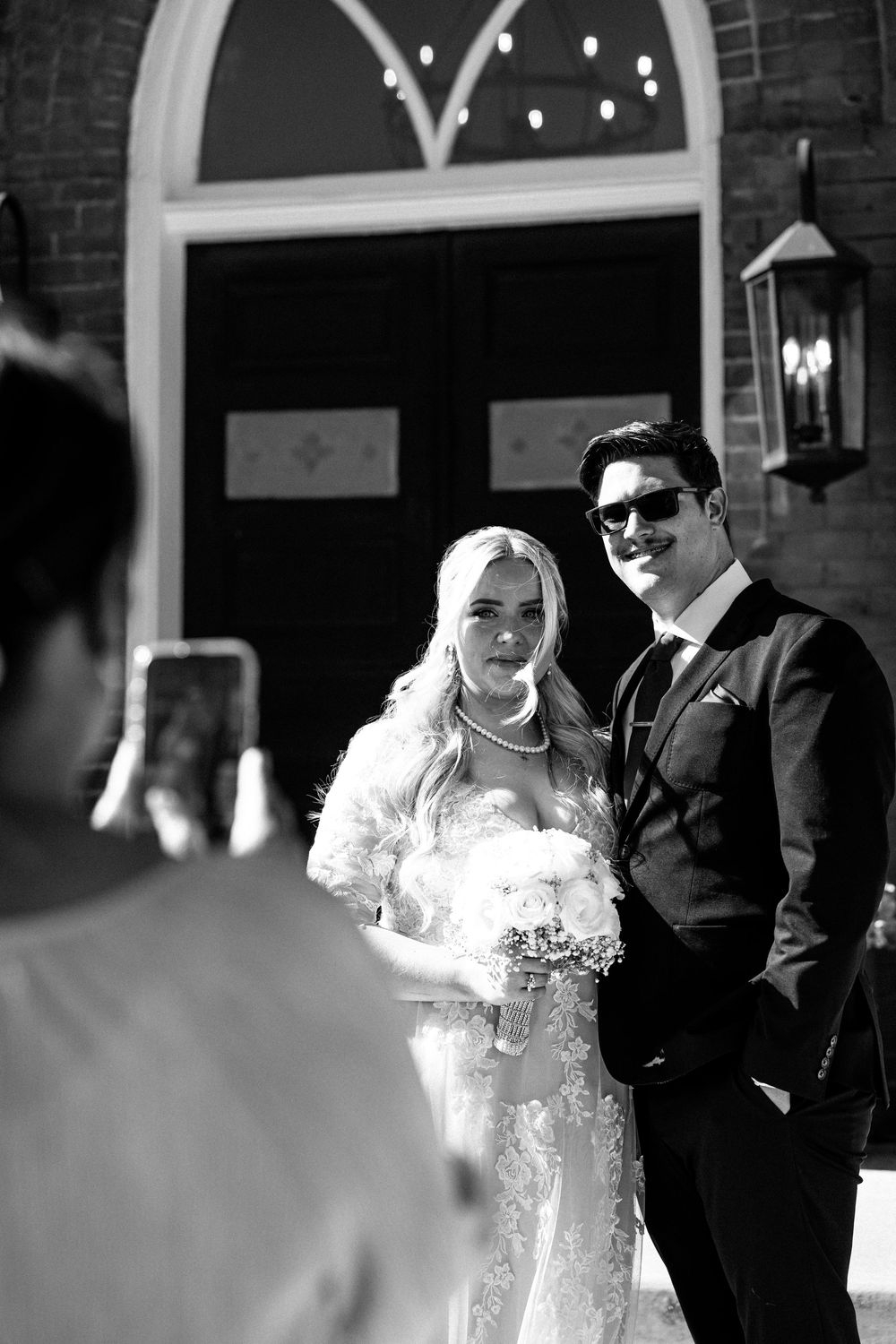 Black and white photo of a bride and groom posing outside a church while a guest photographs them on a smartphone.