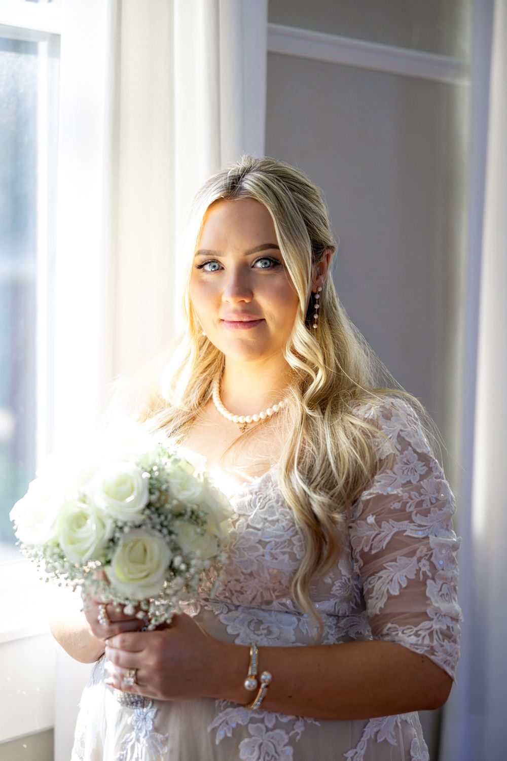 Blonde bride in lace wedding dress holding white rose bouquet, wearing pearl jewelry, standing by window.