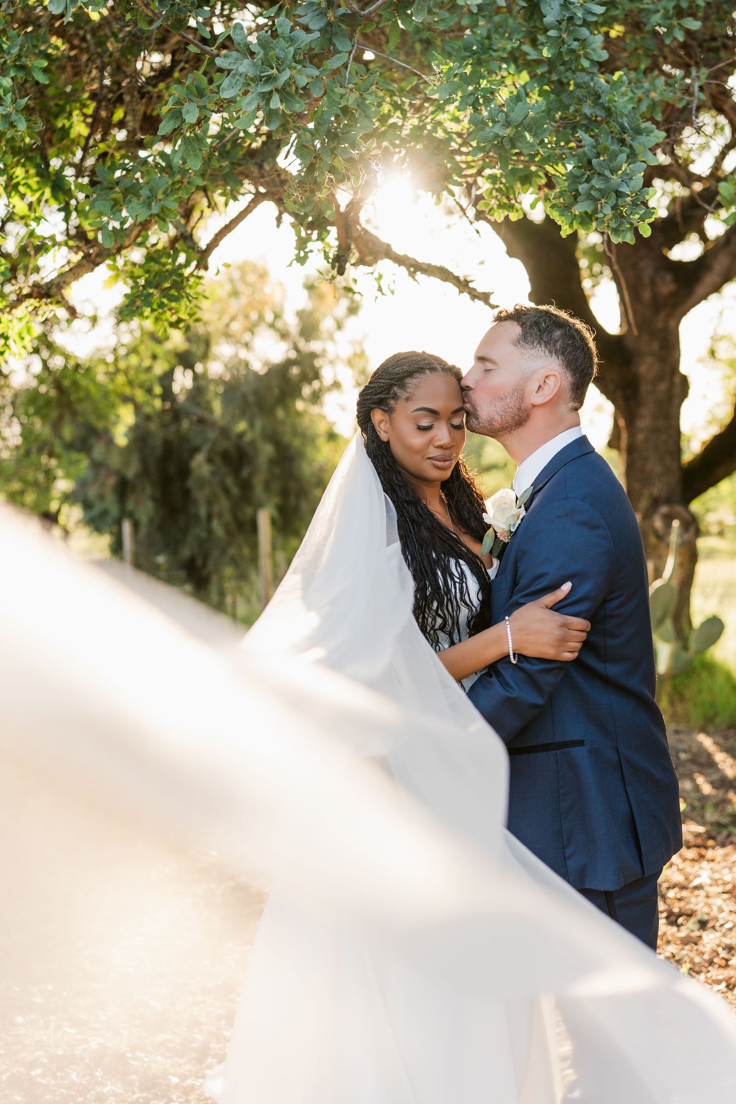 Newlyweds share romantic moments in a sunlit garden setting with trees and flowing white dress details.