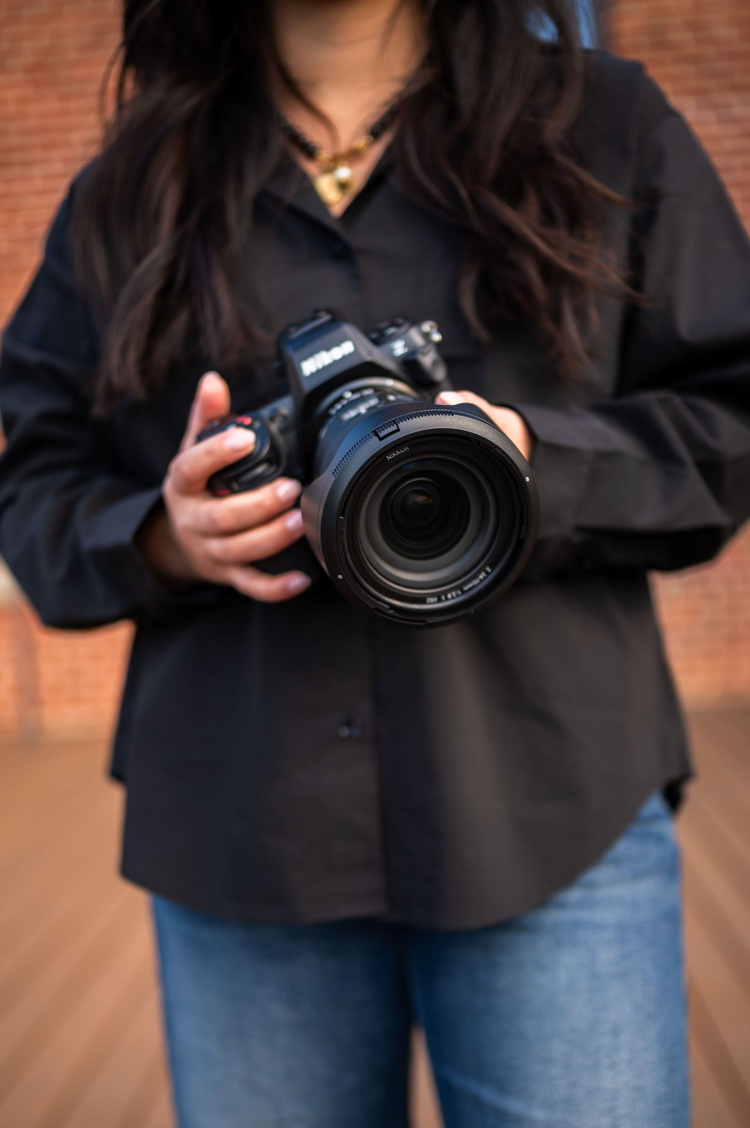 A photographer wearing a black button-down shirt and jeans holds a professional DSLR camera with a large zoom lens.