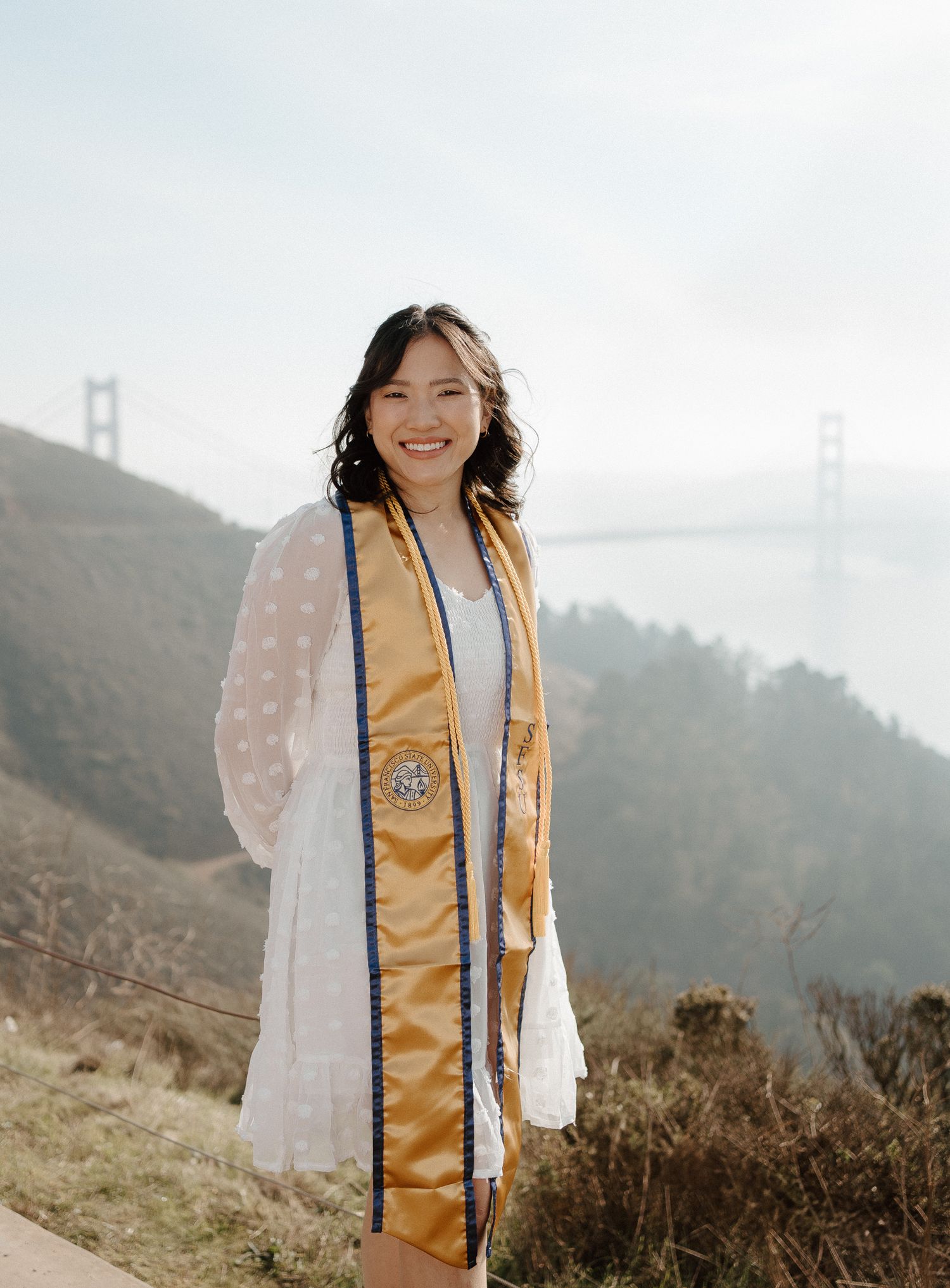 Smiling graduate wearing gold stole poses with Golden Gate Bridge backdrop in San Francisco.