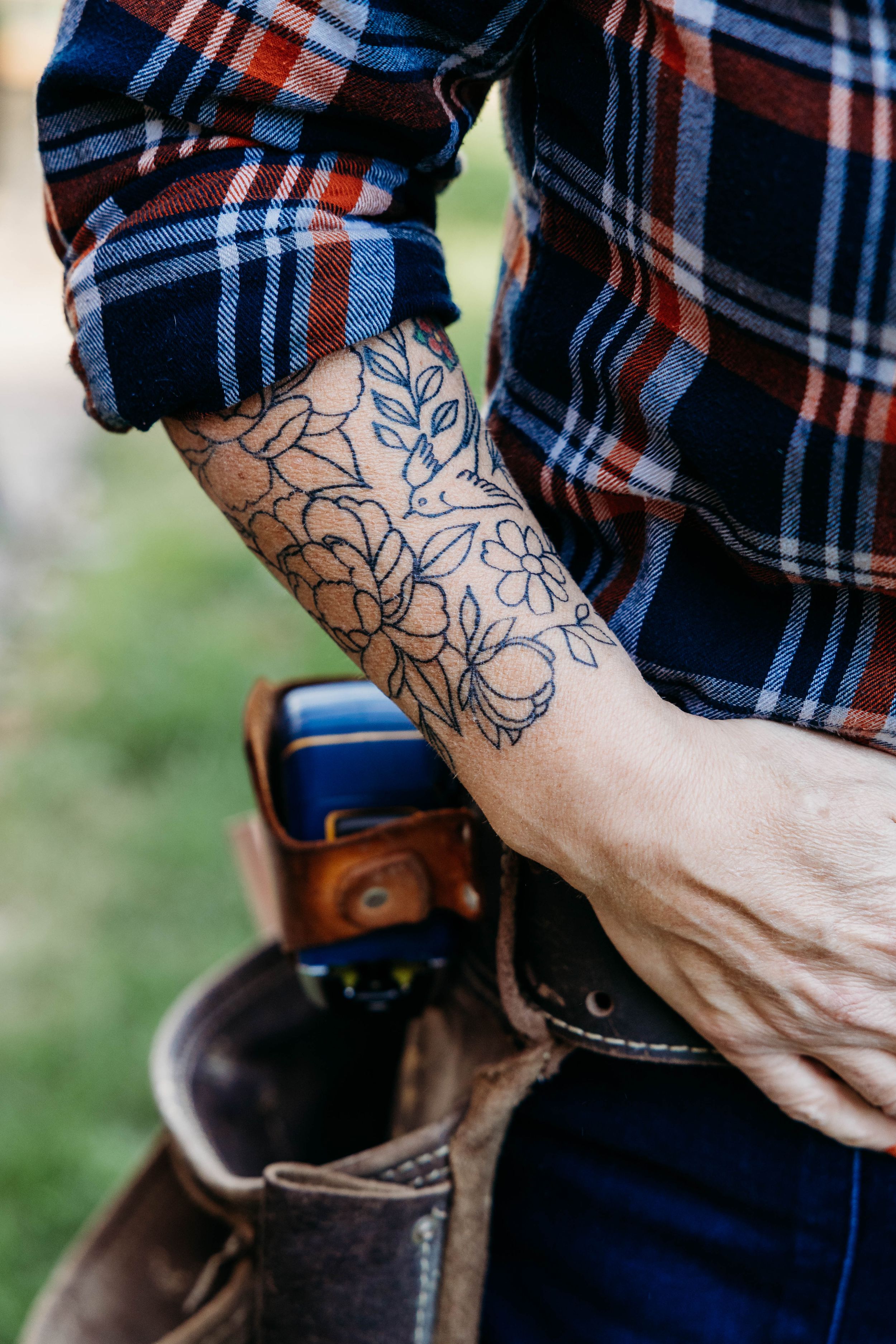Close-up of floral line art tattoo on forearm with plaid shirt sleeve and leather boots in background.