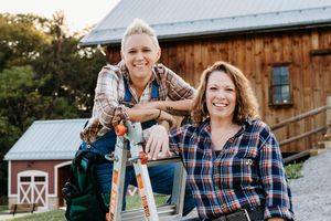 Two people in plaid shirts smile while posing near a wooden barn during golden hour.