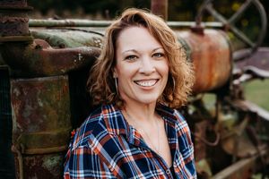 A person in a plaid shirt smiles warmly while posing outdoors against a rustic background.