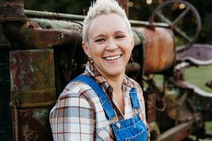 A person wearing denim overalls and plaid shirt poses outdoors near rustic farm equipment and brick walls.