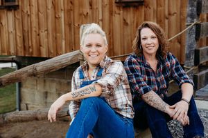 Two people in plaid shirts and jeans sit casually on a wooden fence outside a rustic barn structure.