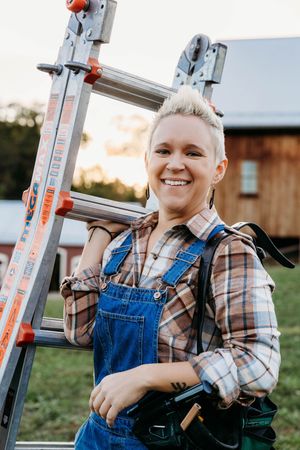 A person in denim overalls and plaid shirt works on a ladder outdoors during sunset.
