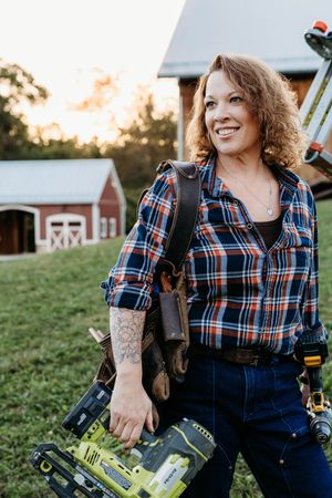 A worker in a plaid shirt and jeans carries tools while standing in front of a red barn at sunset.