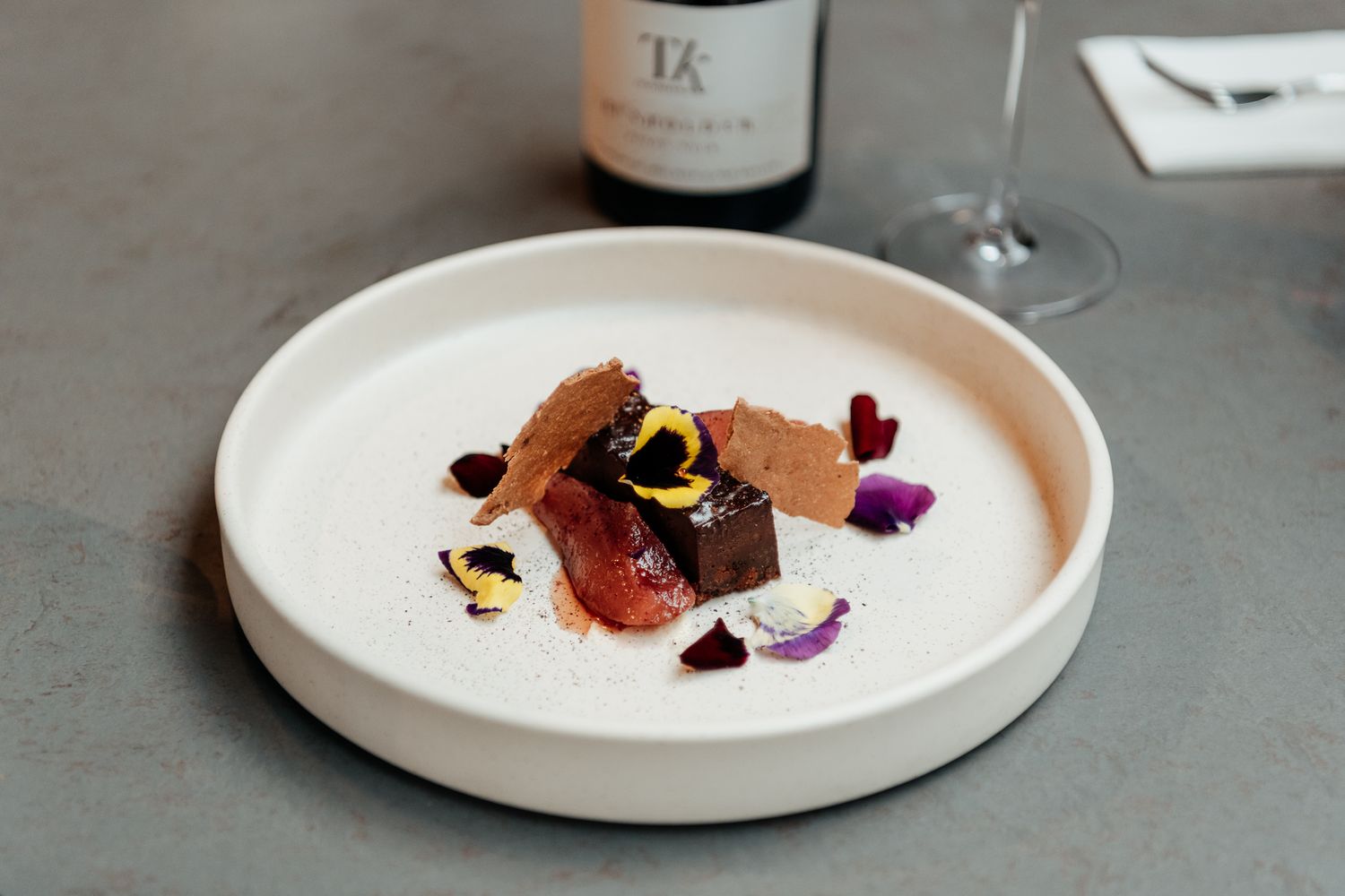 Elegant plated dessert with chocolate and edible flowers on a white ceramic dish against a gray table at The Runholder by August and Vine