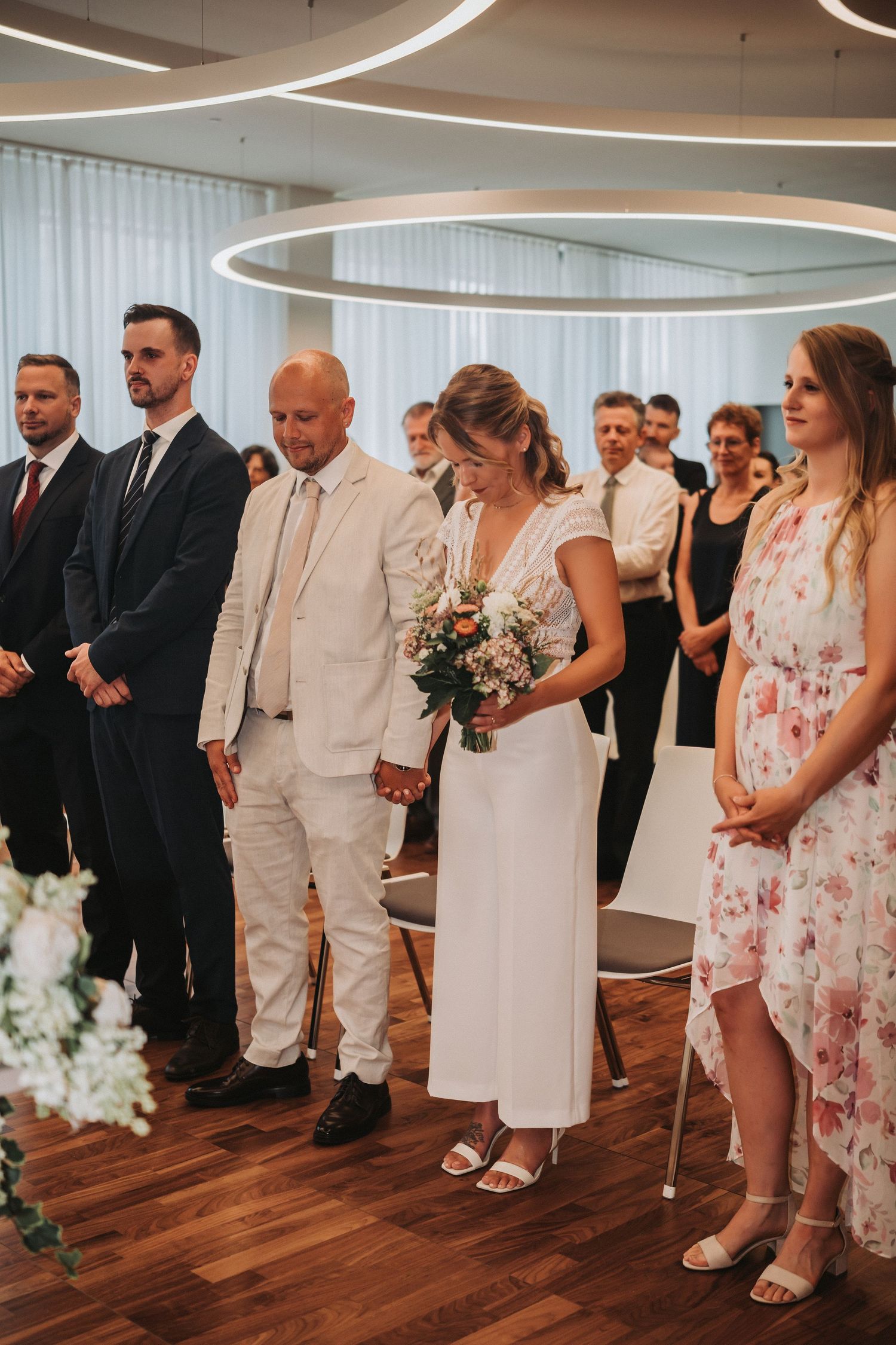 Wedding party lines up in formal attire on wooden floor during indoor ceremony with curved lighting above.