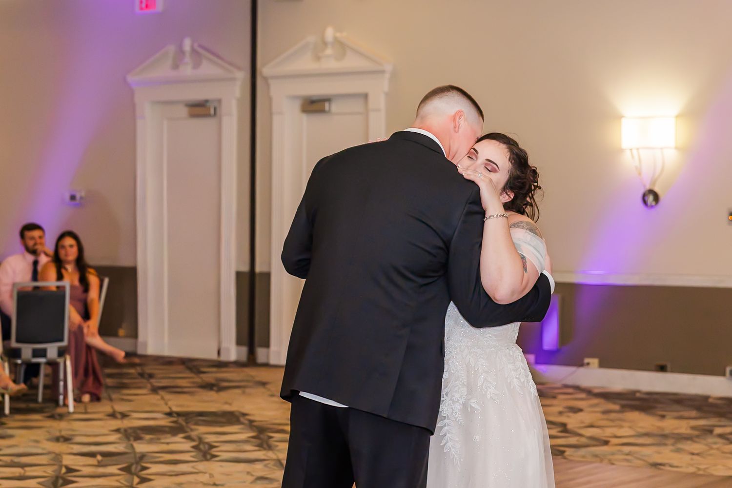 Guests dance and celebrate at an elegant wedding reception in a ballroom with purple uplighting and white decor.